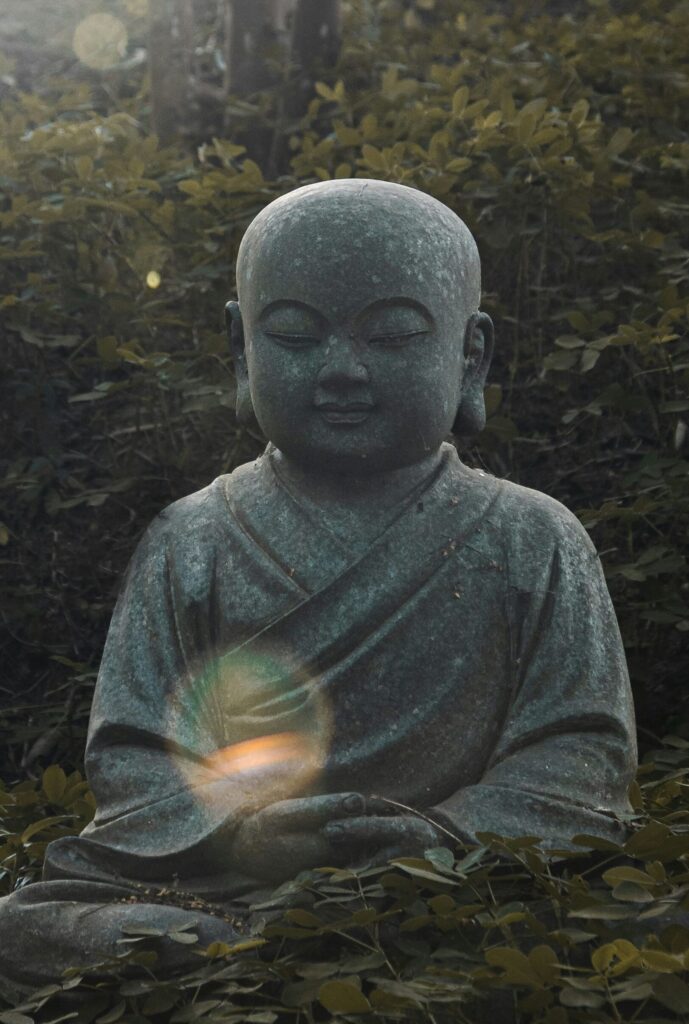 Foto: Kleine graue Buddha-Steinfigur auf grünem Waldboden, ein kreisförmiger Sonnenfleck fällt auf den Oberkörper der Figur. Photo by Gabriel Ramos on Unsplash